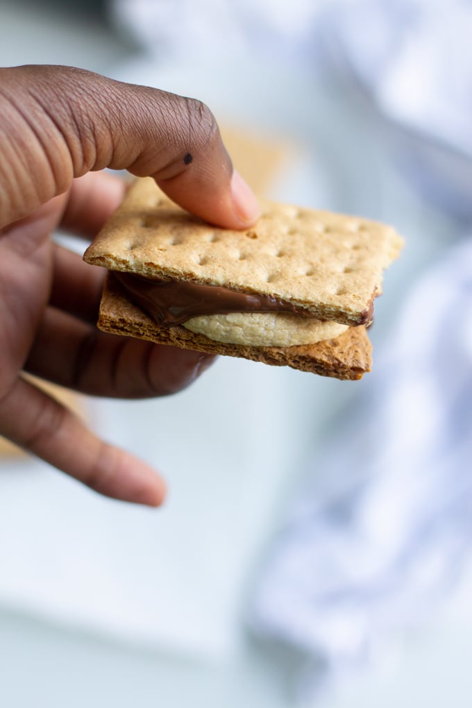You won’t need a campfire to make these s’mores, all you need is an Air Fryer. Delicious roasted marshmallows roasted right in the Air Fryer in less than 10 minutes. It’s the perfect easy dessert. #smallbatchdessert #airfryer #airfryerrecipes #airfryerrecipesfordessert #dessertrecipes hand holding air fryer s'more