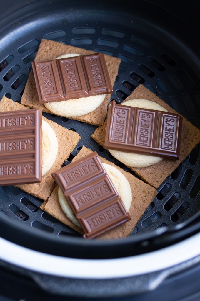 You won’t need a campfire to make these s’mores, all you need is an Air Fryer. Delicious roasted marshmallows roasted right in the Air Fryer in less than 10 minutes. It’s the perfect easy dessert. #smallbatchdessert #airfryer #airfryerrecipes #airfryerrecipesfordessert #dessertrecipes #dessertfortwo graham cracker with marshmallow and chocolate on top in air fryer basket