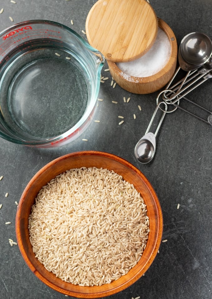 brown rice, salt box, measuring cup with water, and measuring spoons laid out on a gray background