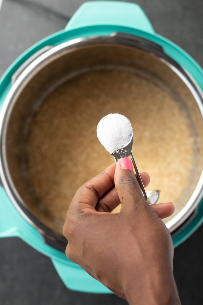 hand holding salt in a measuring spoon over the brown rice and water in the instant pot