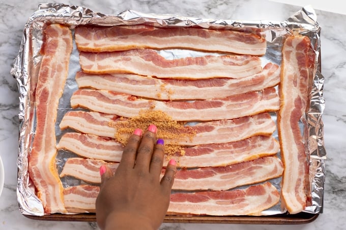 bacon laid on baking pan with foil with hand putting brown sugar in it