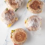 breakfast sandwiches wrapped in plastic wrap on marble with one sandwich unwrapped