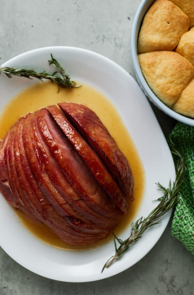 Overhead shot of a glazed holiday ham served on a white plate next to dinner rolls.