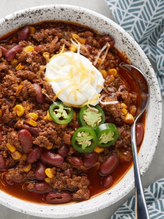 A spoon in a bowl of chili topped with sour cream, shredded cheese and fresh jalapenos.