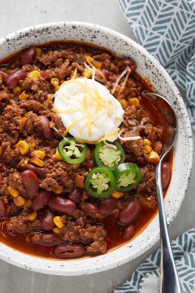 A spoon in a bowl of chili topped with sour cream, shredded cheese and fresh jalapenos.