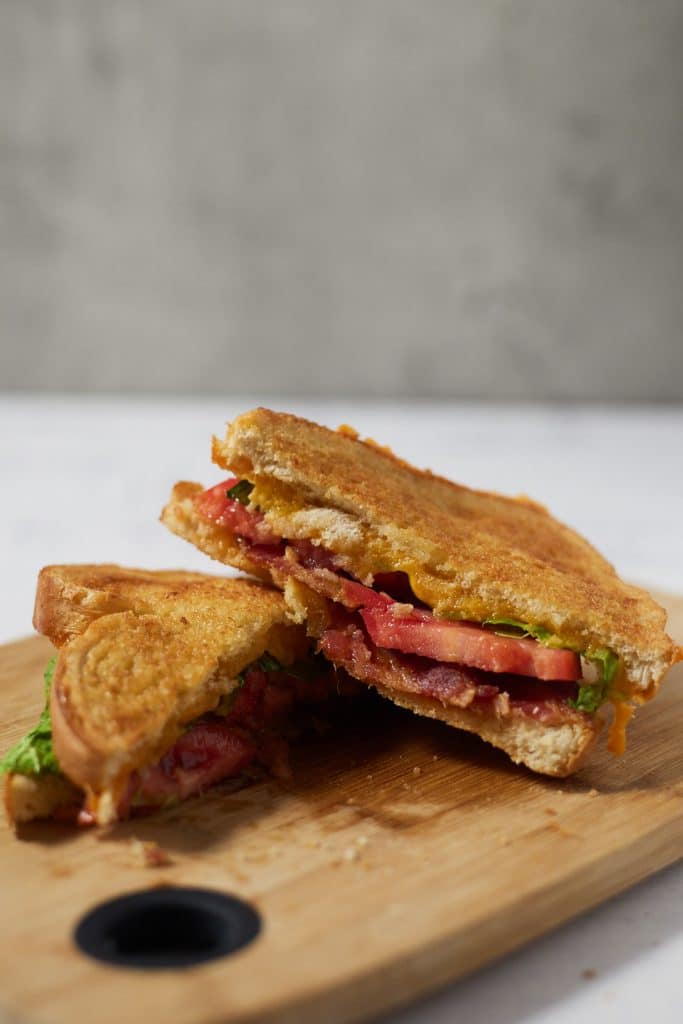 A BLT grilled cheese cut in half on a wooden chopping board.