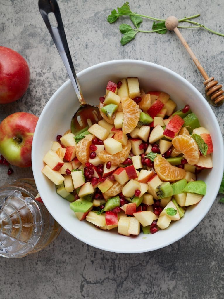 winter fruit salad in a white bowl with a honey pot and fruit surrounding it