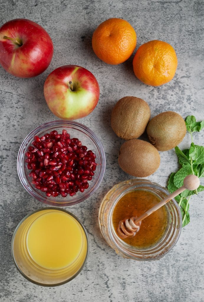 ingredient photo of apples, clementines, kiwi, promegranate seeds, honey, orange juice, and mint leaves