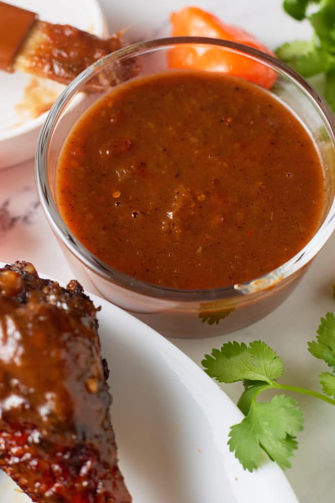 A bowl of jerk bbq sauce next to cooked chicken and fresh herbs.