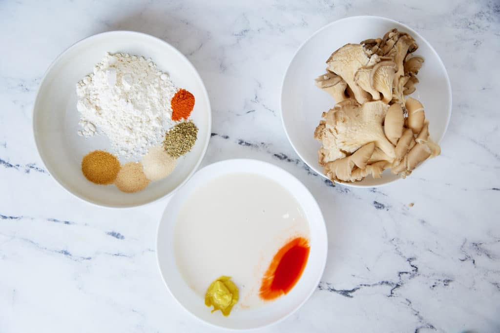 Ingredients to make the recipe on white plates. Flour with seasoning, oyster mushrooms, and almond milk with mustard and hot sauce.