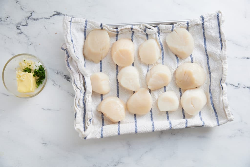Scallops on a baking sheet next to the garlic herb butter mix.