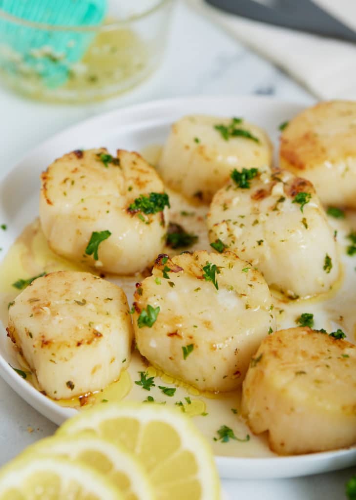 Cooked scallops on a white plate next to slices of lemon.