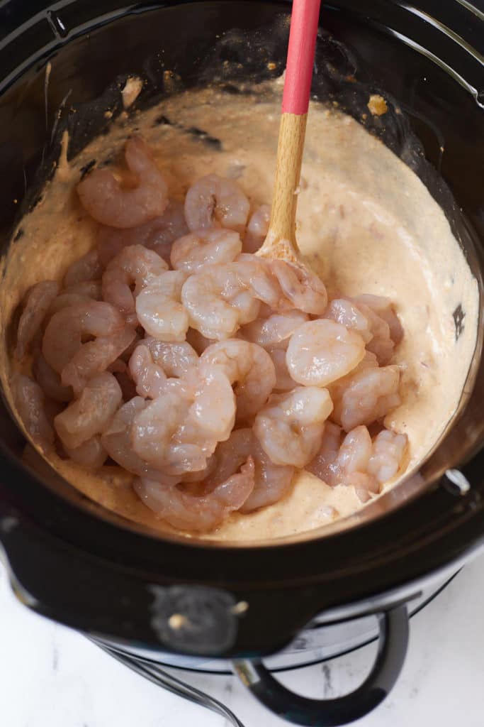 raw shrimp on top of melted cheese mixture in slow cooker with spoon sticking out
