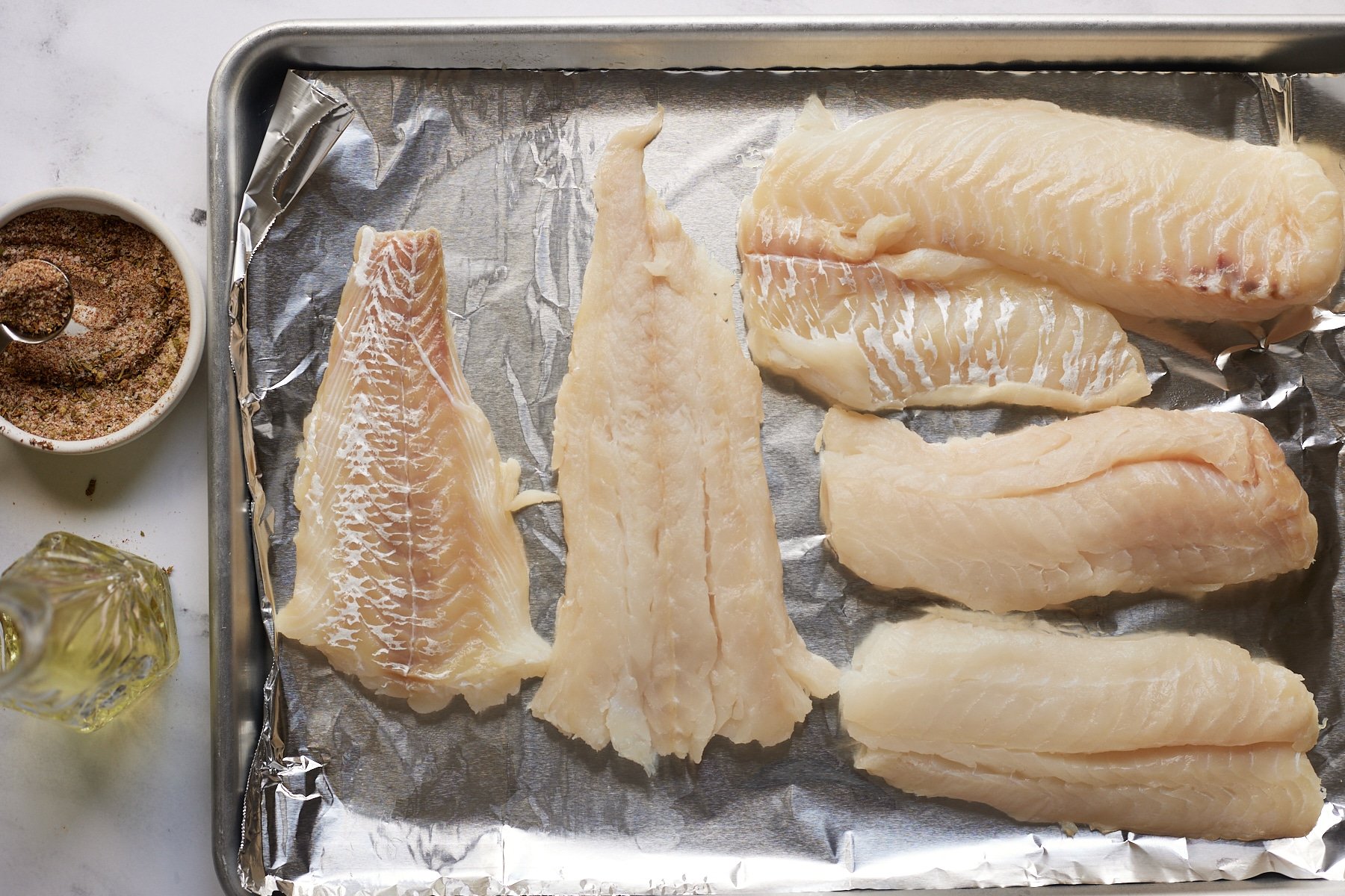 Fish fillets on a lined baking tray next to seasoning and oil.