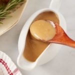 The dripping gravy in a gravy boat with a wooden spoon.