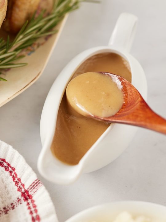 The dripping gravy in a gravy boat with a wooden spoon.