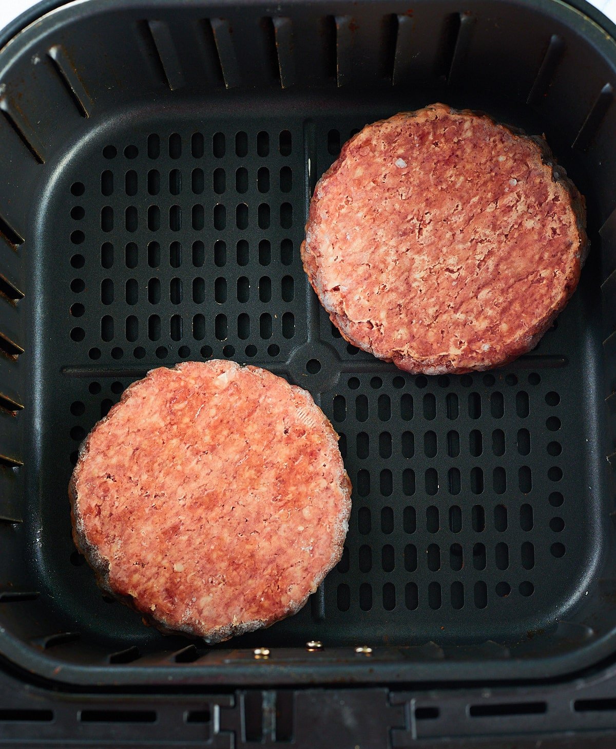 Two frozen hamburger patties in the air fryer basket.