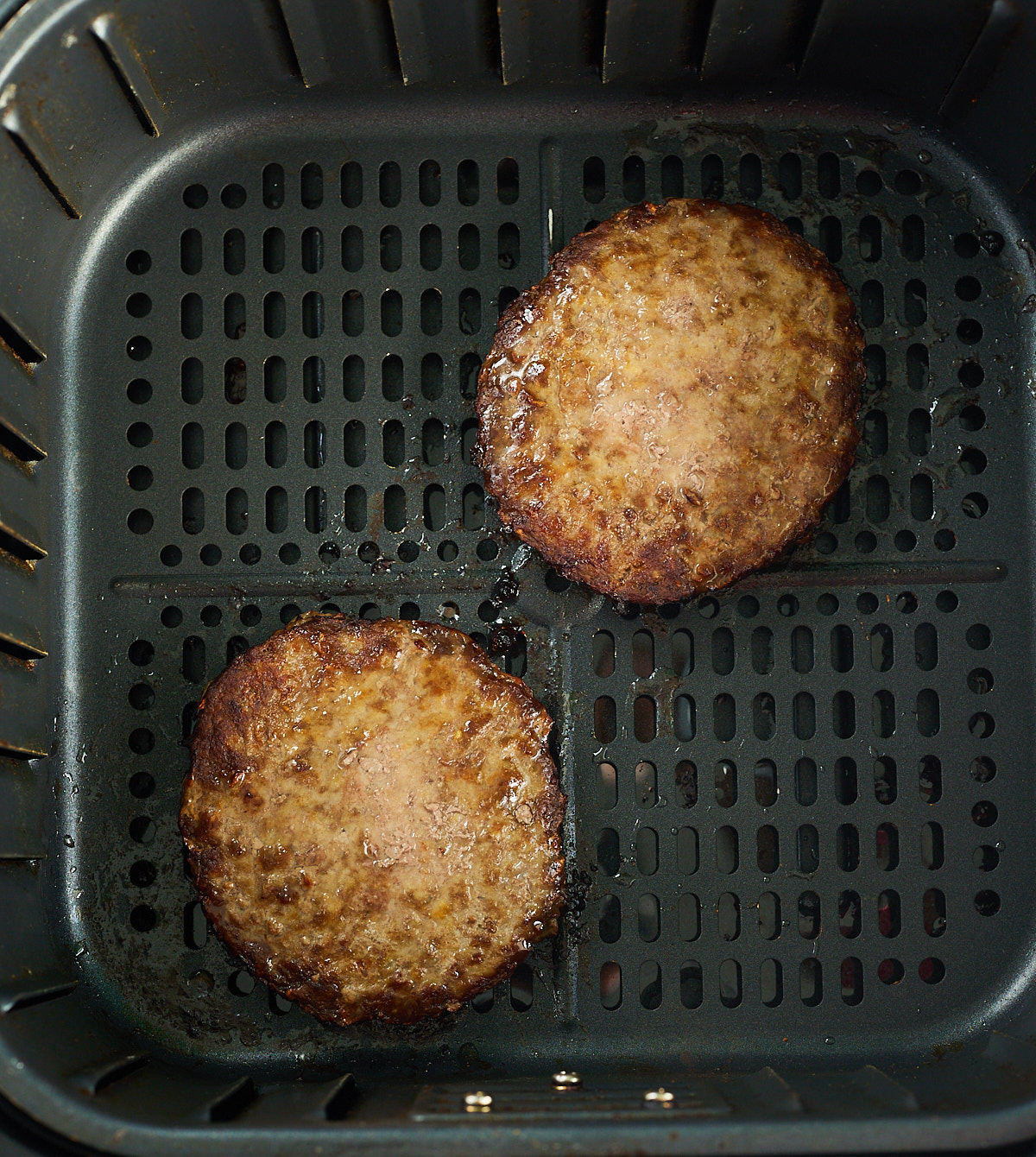 Two cooked hamburger patties in an air fryer basket.