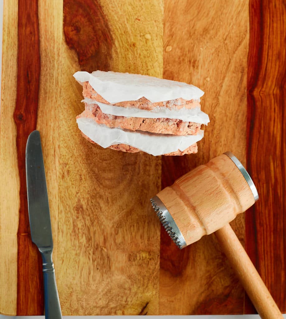 Frozen hamburger patties on a chopping board next to a knife and a mallet.