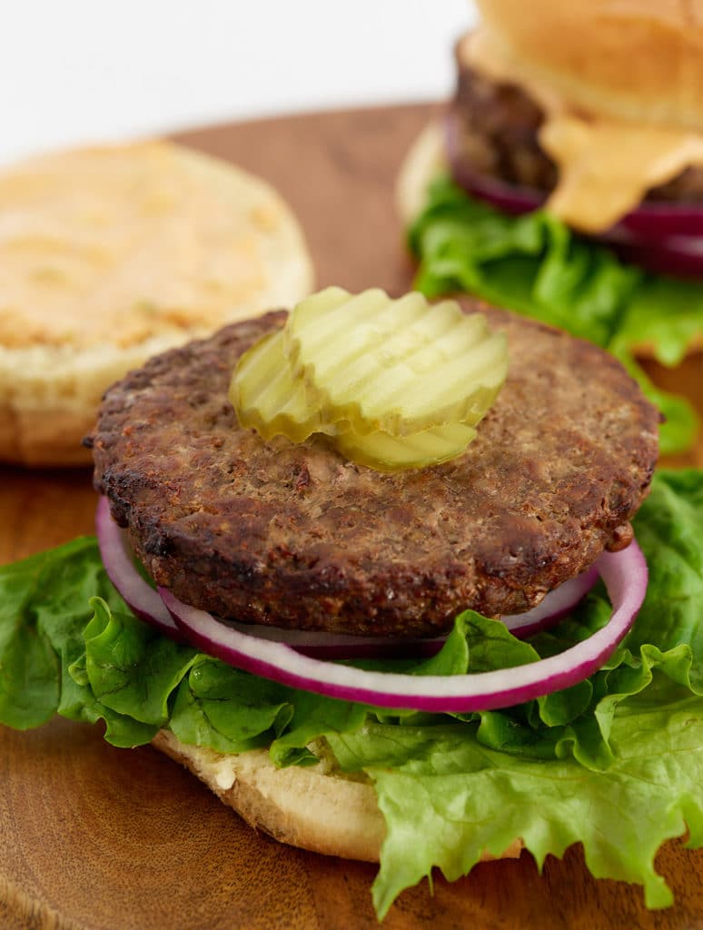 A burger patty sitting on a bun with lettuce, red onion and pickles.