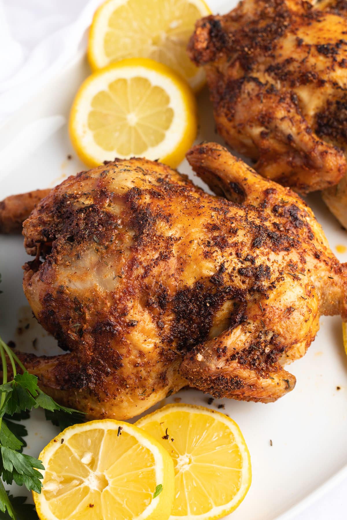 Two cornish hens on a white serving plate with fresh lemon slices and herbs.