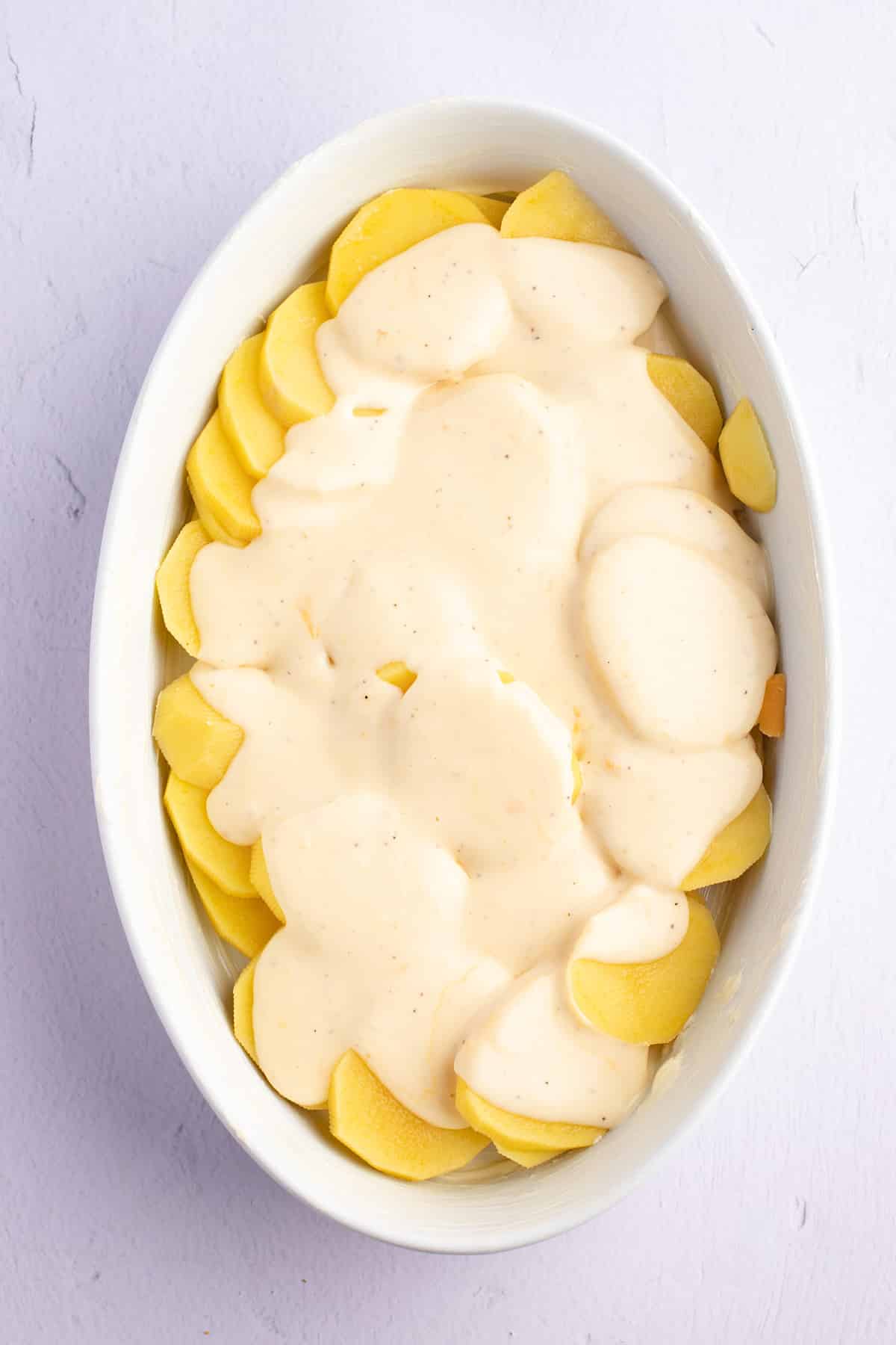 Cream sauce poured over sliced potatoes in a white baking dish.