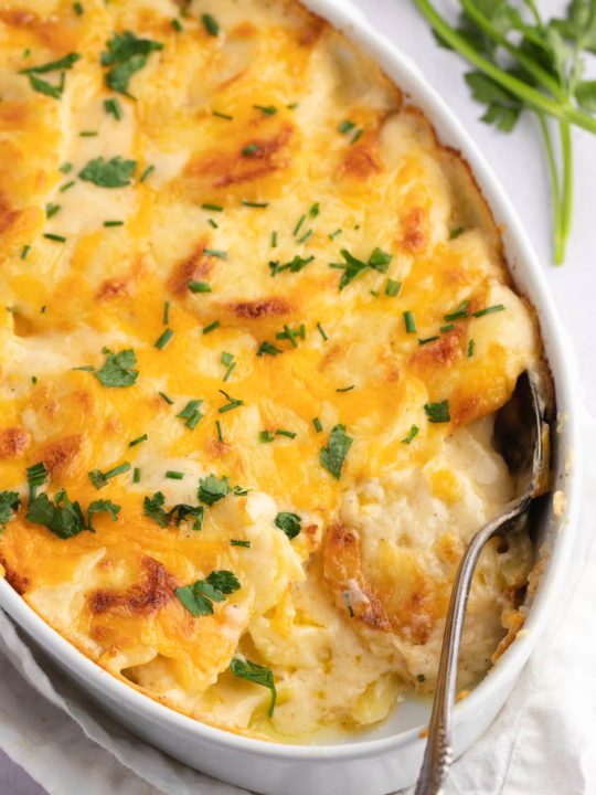 A serving spoon in the baking dish of cheesy scalloped potatoes.