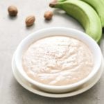 green banana porridge in white bowl with green banana and nutmeg in background