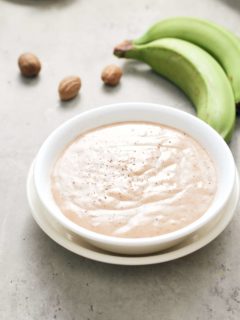 green banana porridge in white bowl with green banana and nutmeg in background