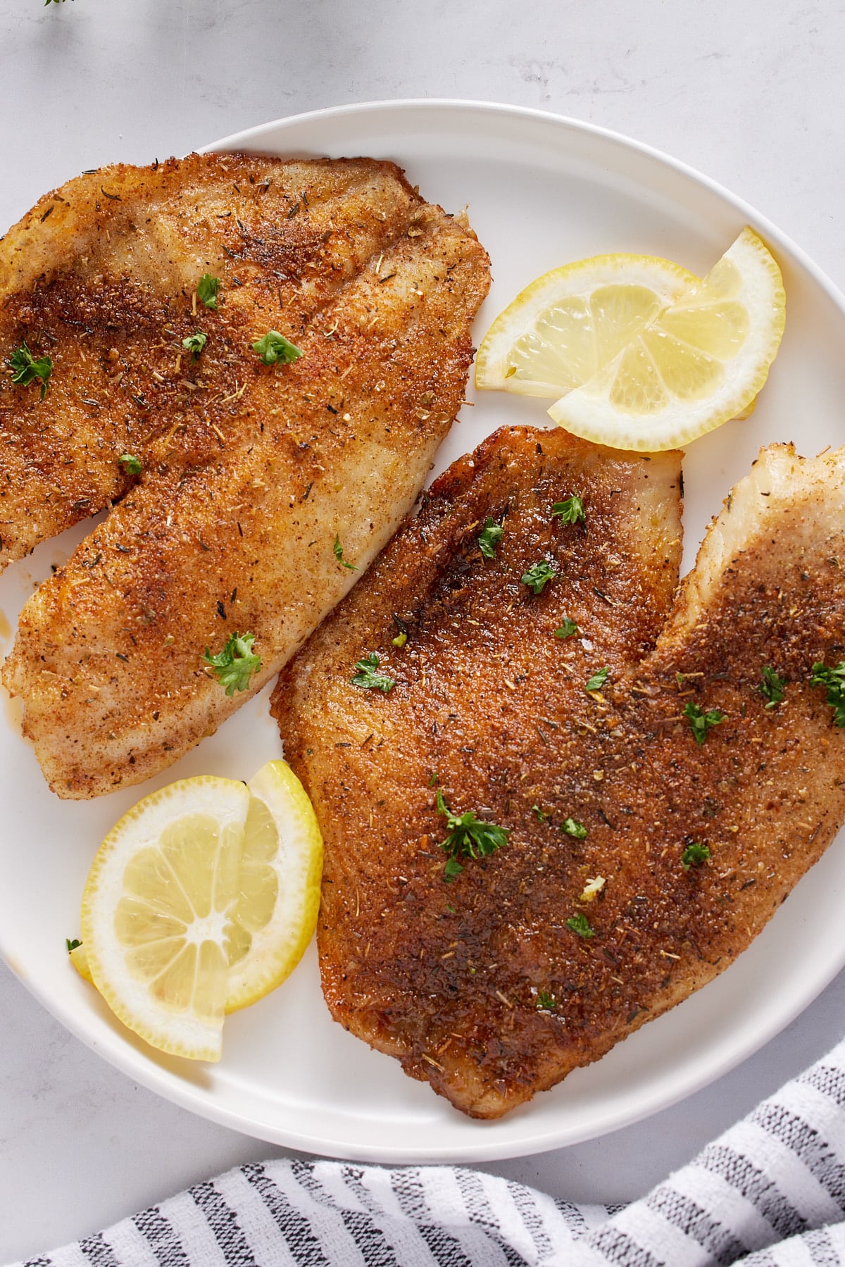 Two air fryer tilapia fillets on a plate with lemon slices.