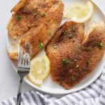 A fork cutting a tilapia fillet on a white plate.