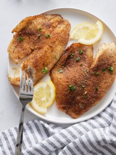 A fork cutting a tilapia fillet on a white plate.