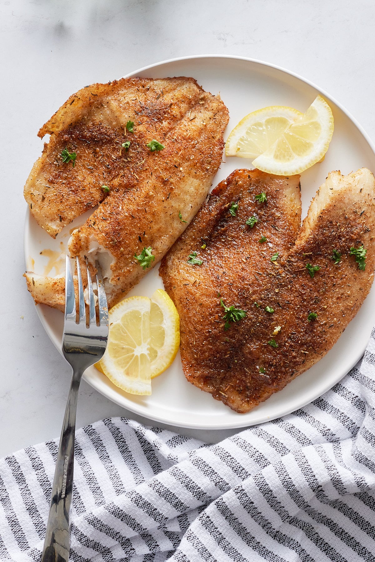 A fork cutting a tilapia fillet on a white plate.