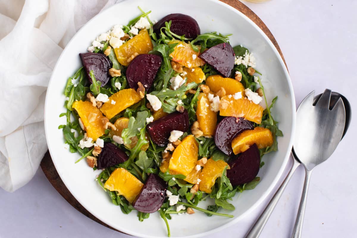 Beet and orange salad in a white bowl next to serving spoons.