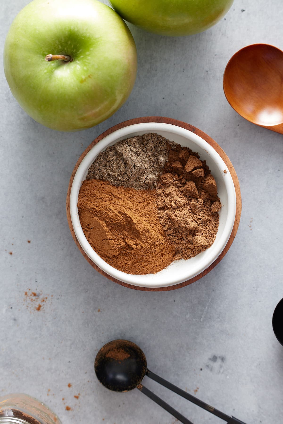 Top down image of green apples, a small white bowl of mixed spices and a teaspoon