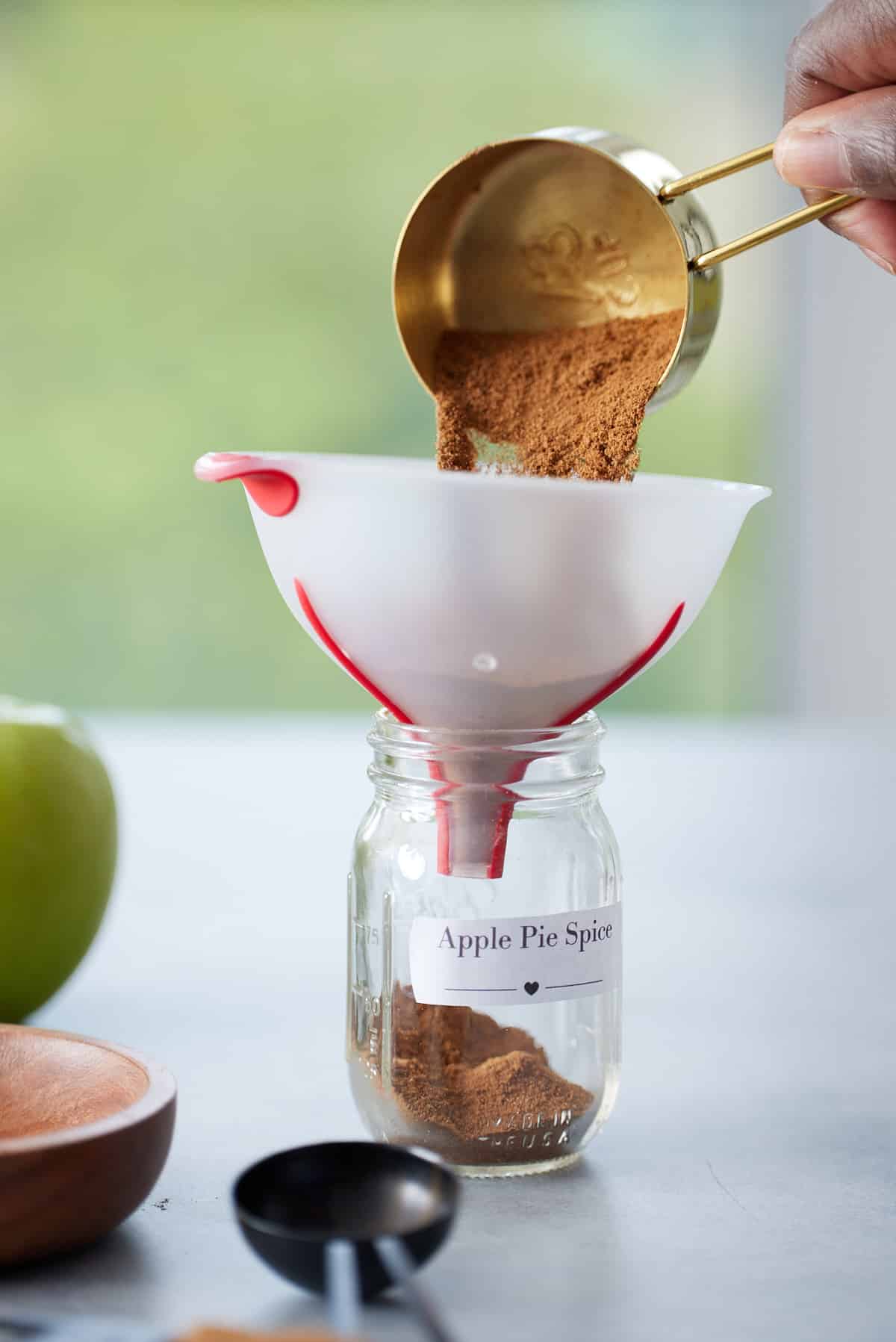 A glass jar with a funnel attached and a cup of spice blend pouring in