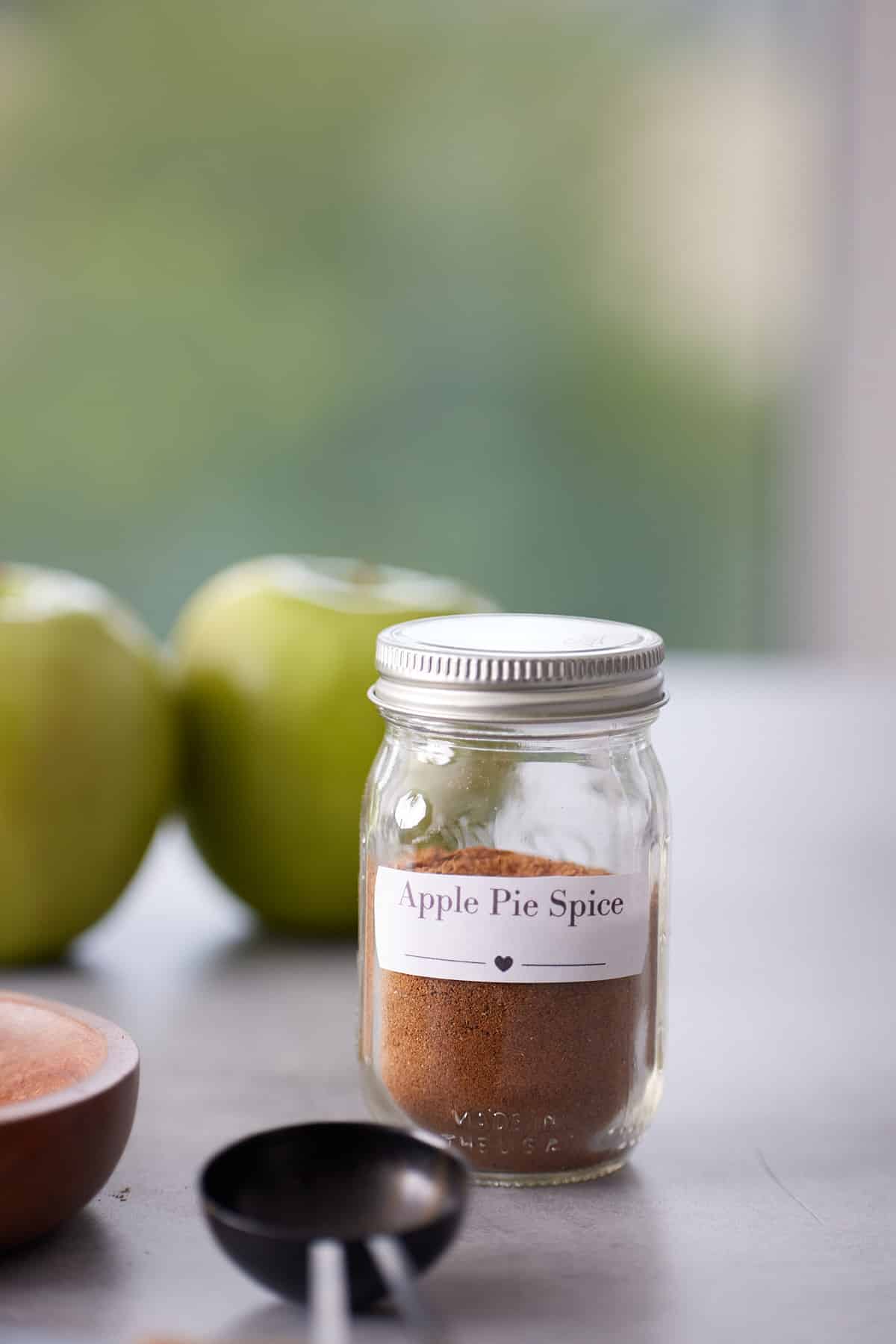 Jar of apple pie spice mix with green apples and a tablespoon