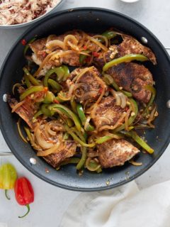 A black skillet containing brown fish stew with a bowl of rice and peas and Scotch bonnet peppers set alongside