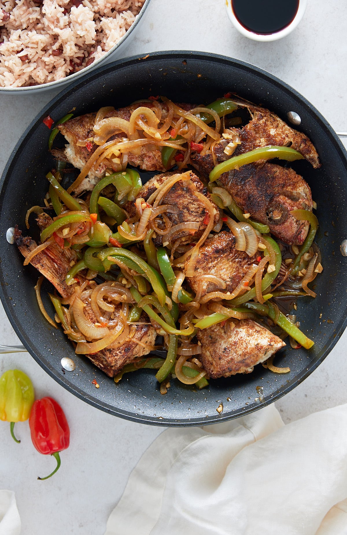 A black skillet containing brown fish stew with a bowl of rice and peas and Scotch bonnet peppers set alongside