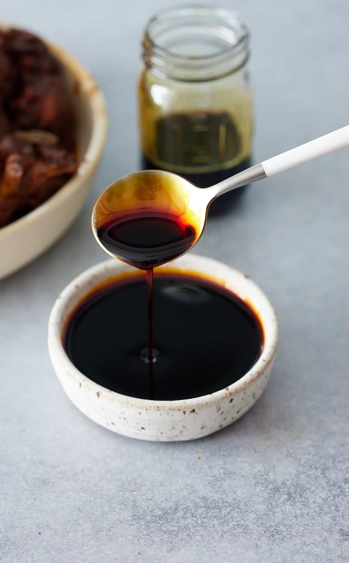 Browning sauce being poured from a spoon into a small white bowl of sauce with a jar of sauce in the background