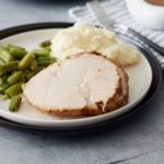 White plate with slices of turkey breast, mashed potato and green beans and a gravy jug, napkin and cutlery set alongside