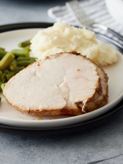 White plate with slices of turkey breast, mashed potato and green beans and a gravy jug, napkin and cutlery set alongside
