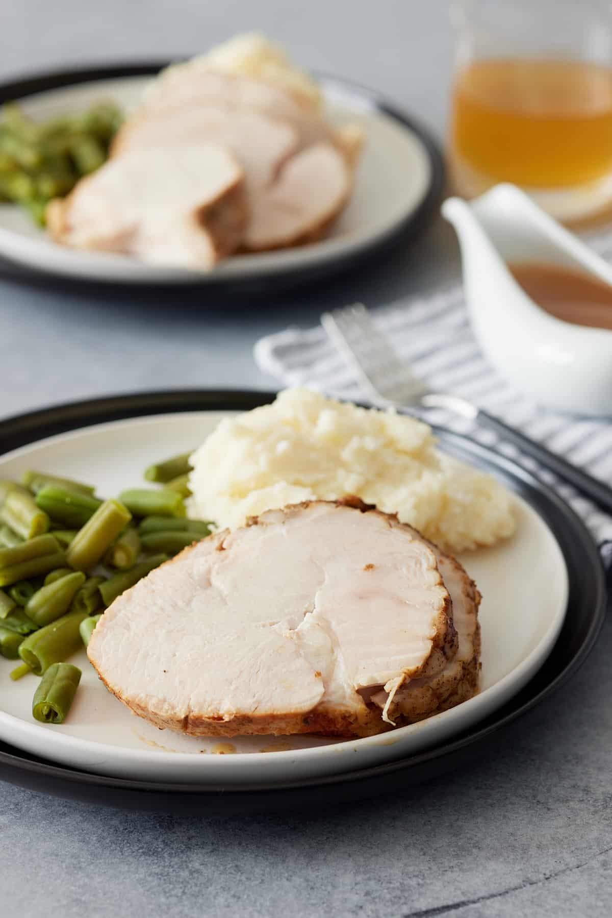 Table set with two servings of turkey breast, mashed potato and green beans set on white plates with a gravy jug set alongside