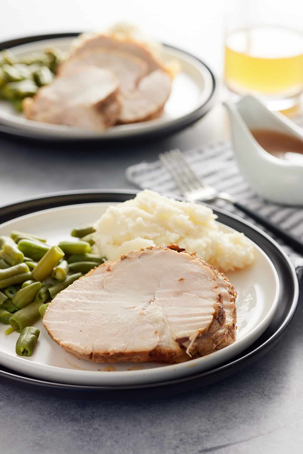 Table set with two servings of turkey breast, mashed potato and green beans set on white plates with a gravy jug set alongside