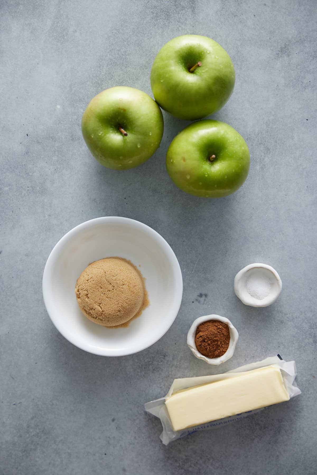 Recipe ingredients for Southern fried apples including 3 green apples and other ingredients set out in bowls on a marble surface
