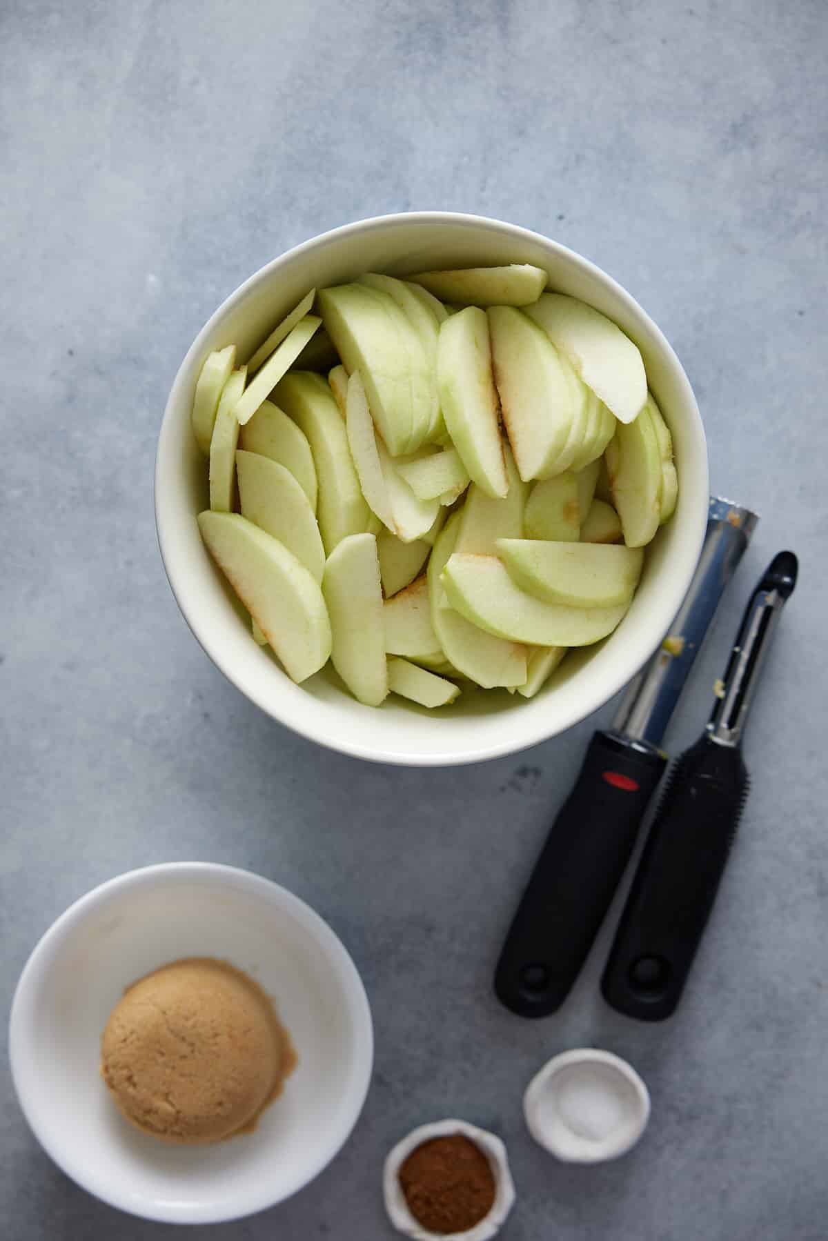 Recipe ingredients for fried apples set out in white bowls on a marble surface with apple peeler and corer set alongside