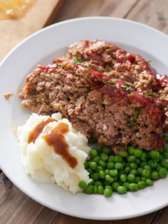 White plate with 2 slices of Southern meatloaf, mash potato, gravy and green peas.
