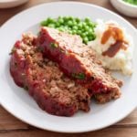 White plate with 2 slices of Southern meatloaf, mash potato, gravy and green peas.