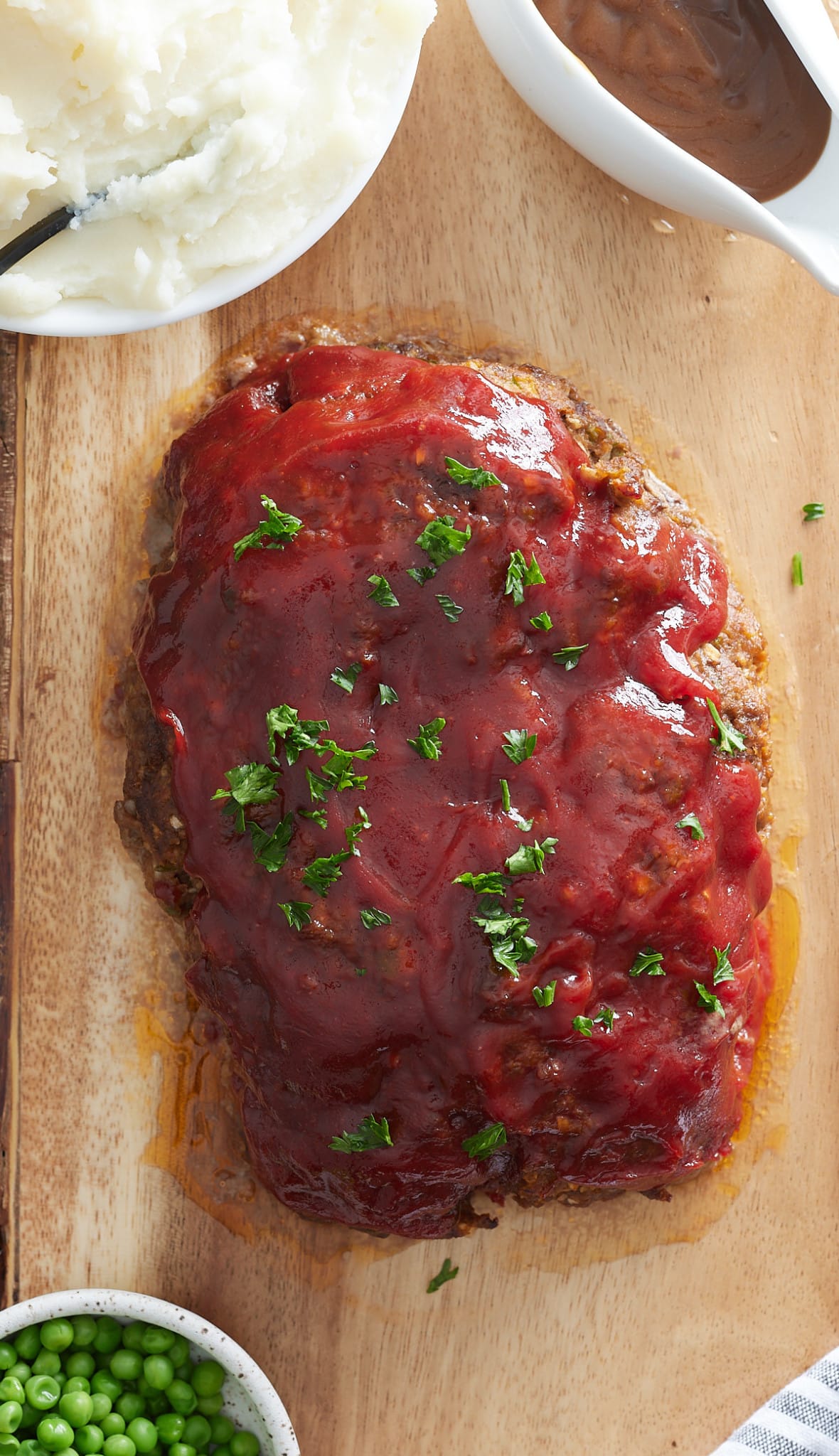 Glazed meatloaf set on a wooden board, with bowl of mash, bowl of green peas and gravy jug set alongside