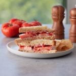 Close up image of a Southern tomato sandwich set on a white plate with some tortilla chips and fresh tomatoes and salt and pepper grinders in background